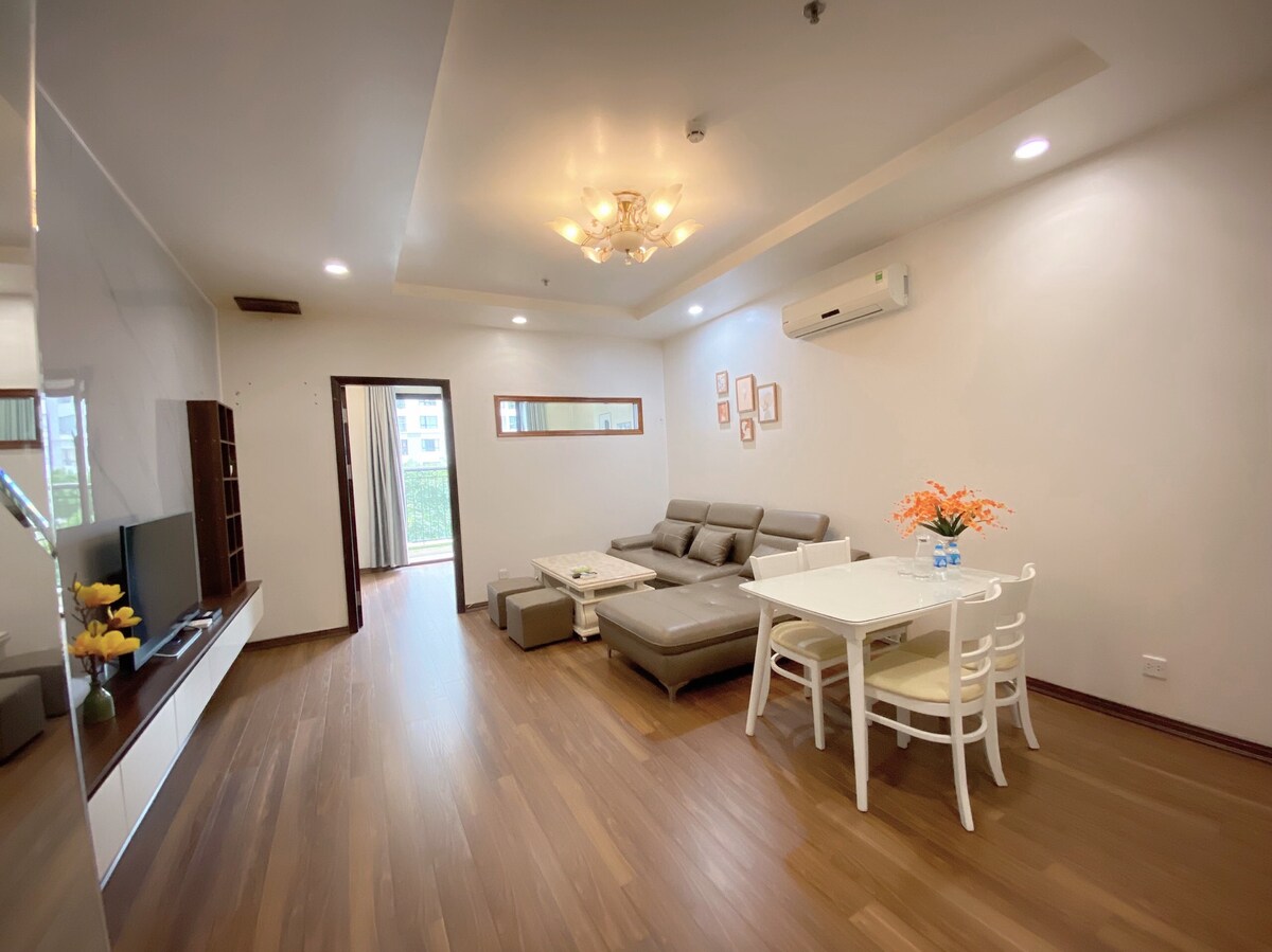 A bright living area features a comfortable sectional sofa with neutral tones and a wooden coffee table. A dining table with white chairs is positioned nearby. Natural light is visible through a window, enhancing the warmth of the space.