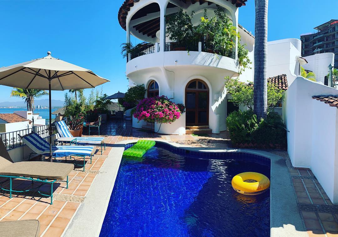 A bright view of the outdoor area showcases a sparkling blue private pool surrounded by lounge chairs. An umbrella provides shade beside the water, while colorful inflatable floats rest on the surface. Lush greenery and vibrant flowers enhance the tropical ambiance of the villa.
