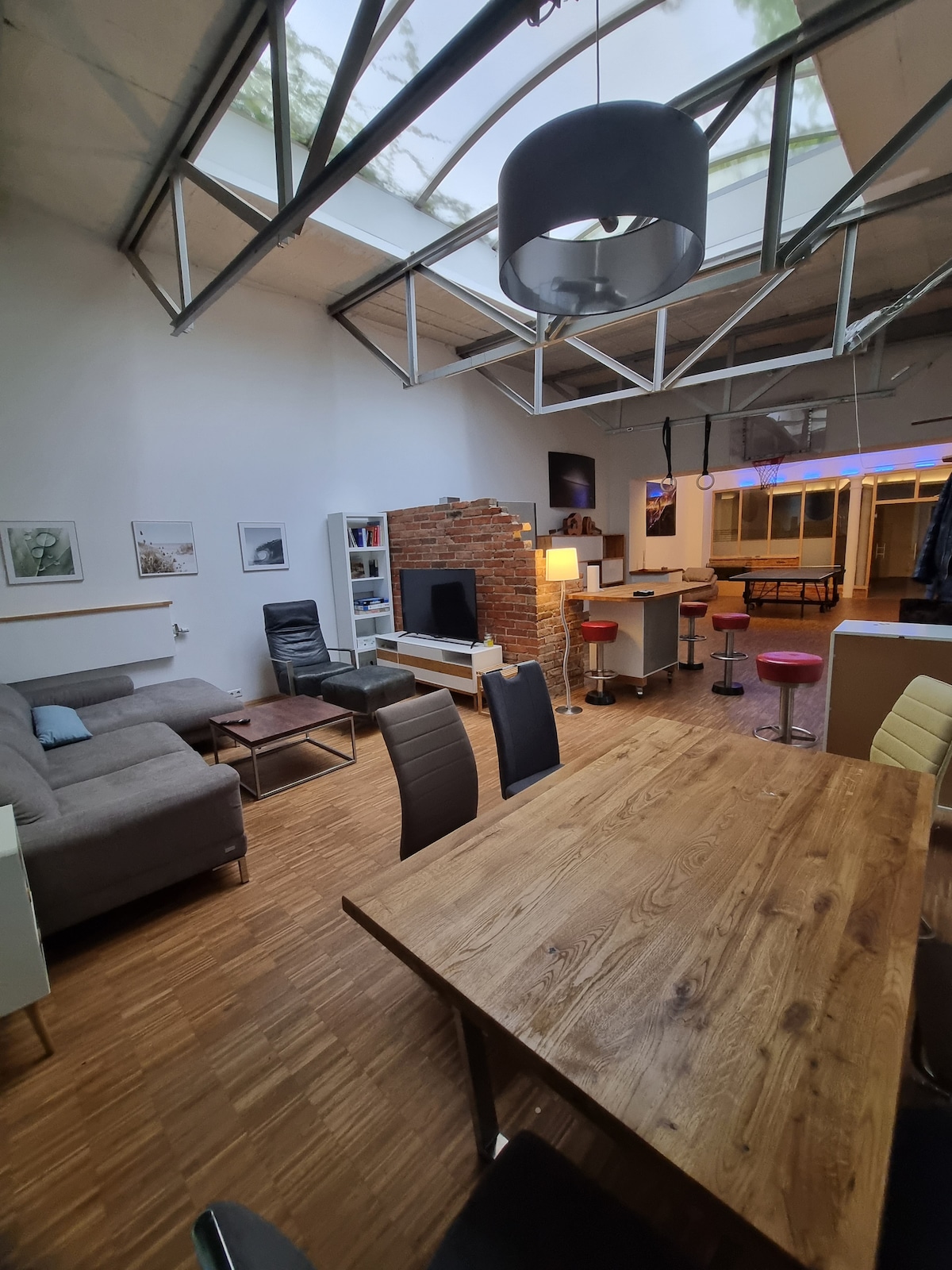 A spacious living area is illuminated by natural light streaming through a glass roof. A comfortable sofa is positioned by exposed brick features, while a wooden dining table is surrounded by upholstered chairs. Bar stools are visible near a sleek counter, adding to the inviting space.