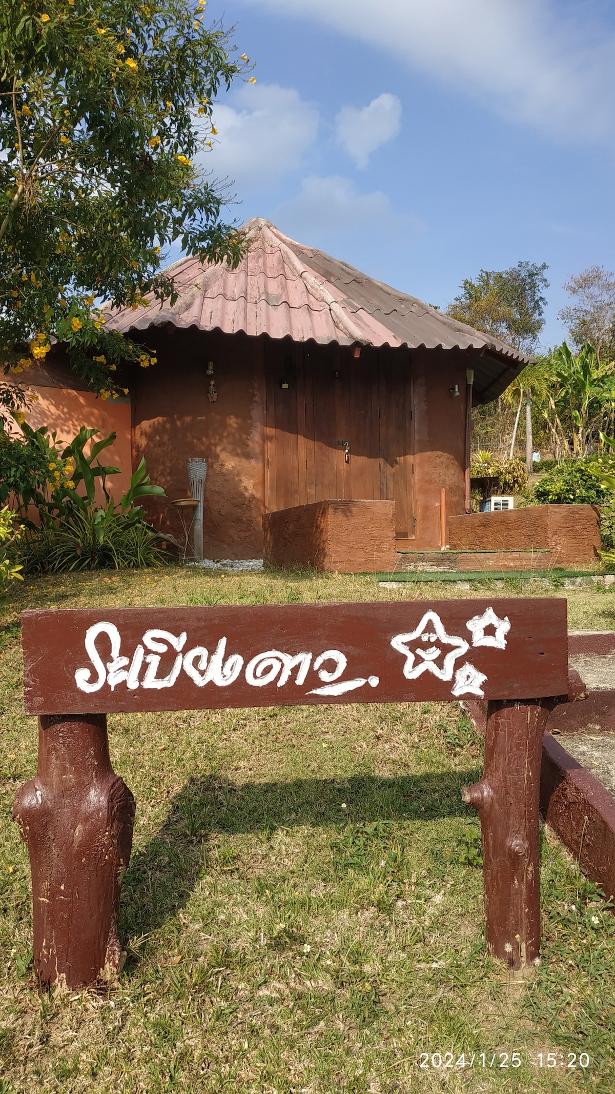 A wooden sign with stylized lettering marks the entrance to a clay structure surrounded by natural greenery. The quaint building features a thatched roof and earthy tones, set against a clear blue sky, creating a harmonious connection with the environment.
