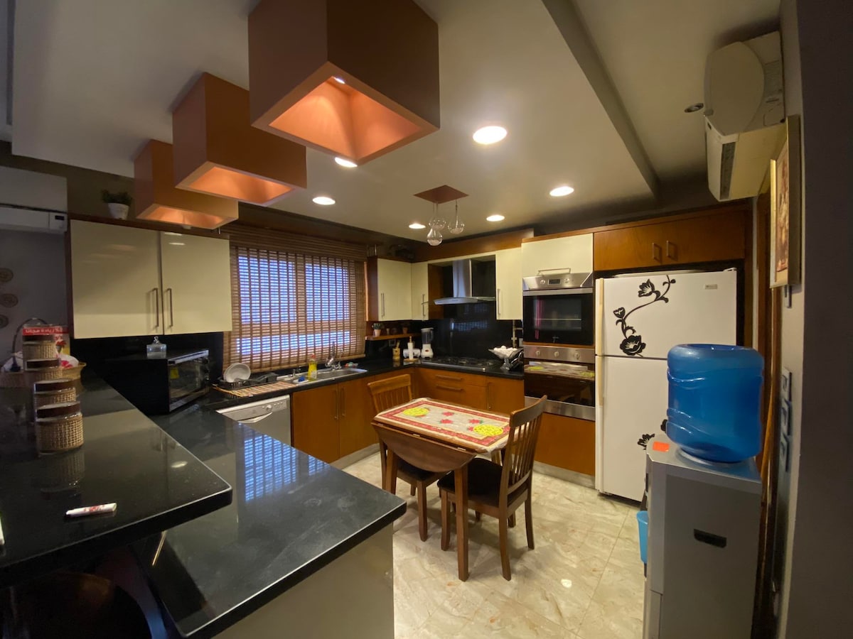 The kitchen features modern cabinetry and sleek black countertops, illuminated by pendant lights. A dining table with wooden chairs is positioned nearby, while kitchen appliances, including a refrigerator and stove, are neatly arranged. Large windows allow natural light to fill the space.