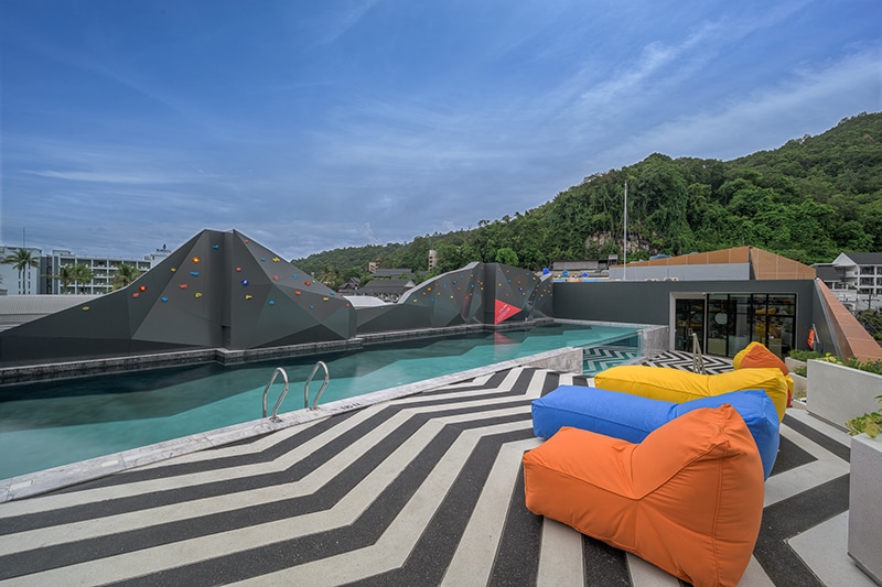 A rooftop area features a swimming pool bordered by vibrant geometric climbing wall designs, providing a modern aesthetic. Colorful bean bags in orange and blue are arranged along a striking striped pattern on the flooring, contrasting with the greenery in the background.