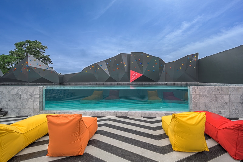 A rooftop swimming pool is visible, framed by colorful bean bags in orange and yellow. Above, a decorative wall features geometric shapes with bright accents, complementing the clear sky and serene water. The area is designed for relaxation and enjoyment.