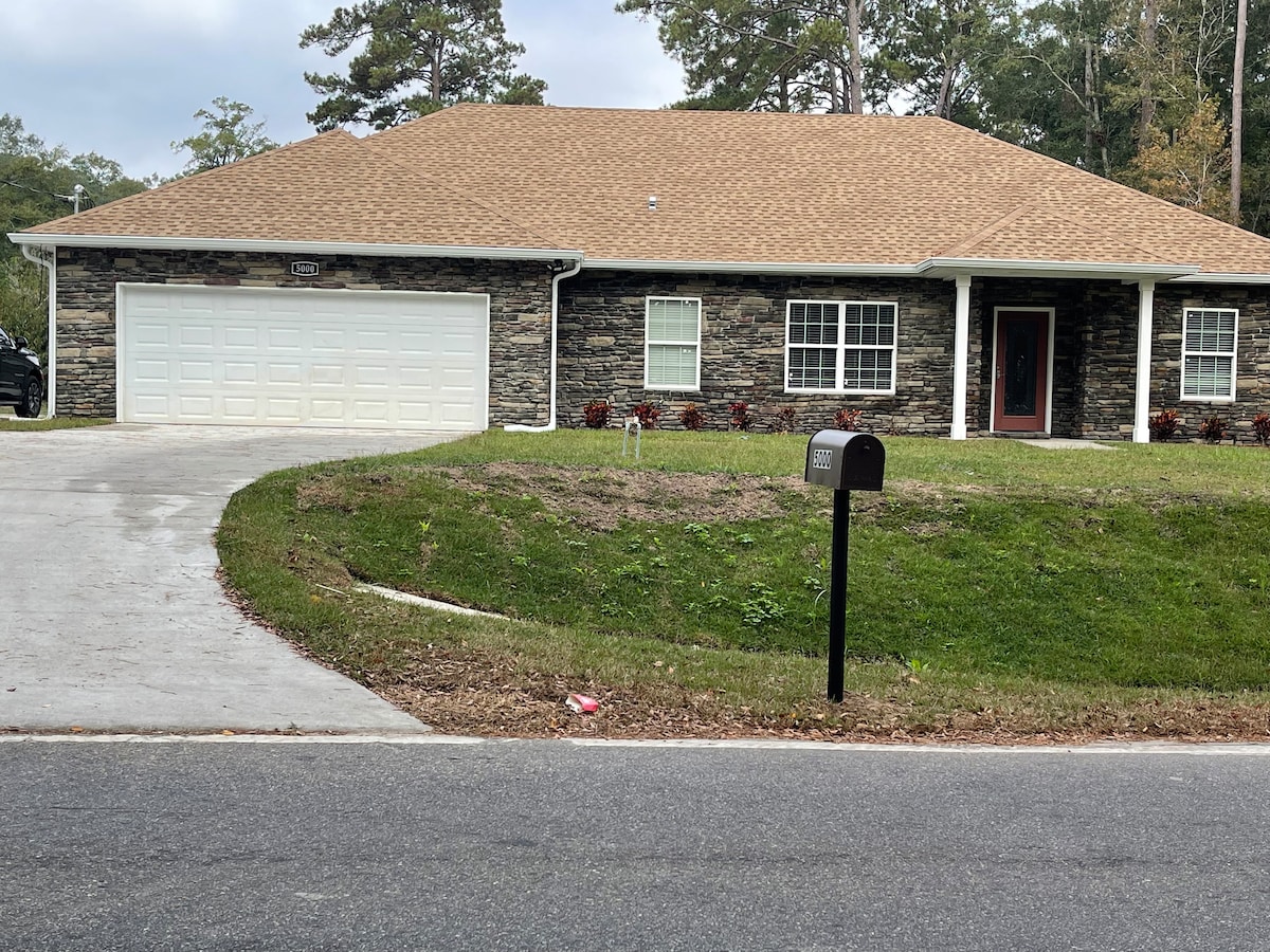 The exterior of a modern single-story home is displayed, featuring a stone facade and a light-colored roof. A spacious driveway leads to a two-car garage. Well-maintained landscaping includes a grassy area and decorative plants near the front entrance, enhancing the welcoming appearance.
