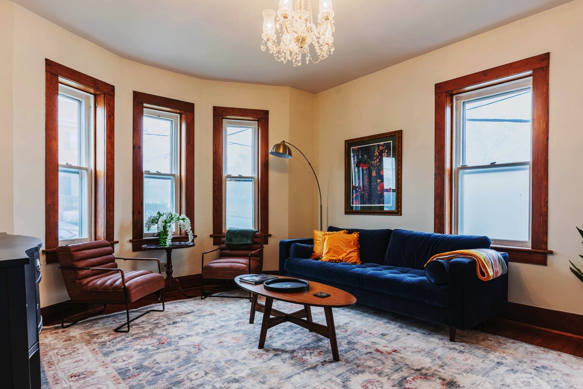 A bright living room features large windows framed in stained wood. A plush velvet couch is complemented by modern leather chairs. A chandelier hangs above, and a patterned area rug covers the hardwood floor, creating a comfortable space for relaxation.
