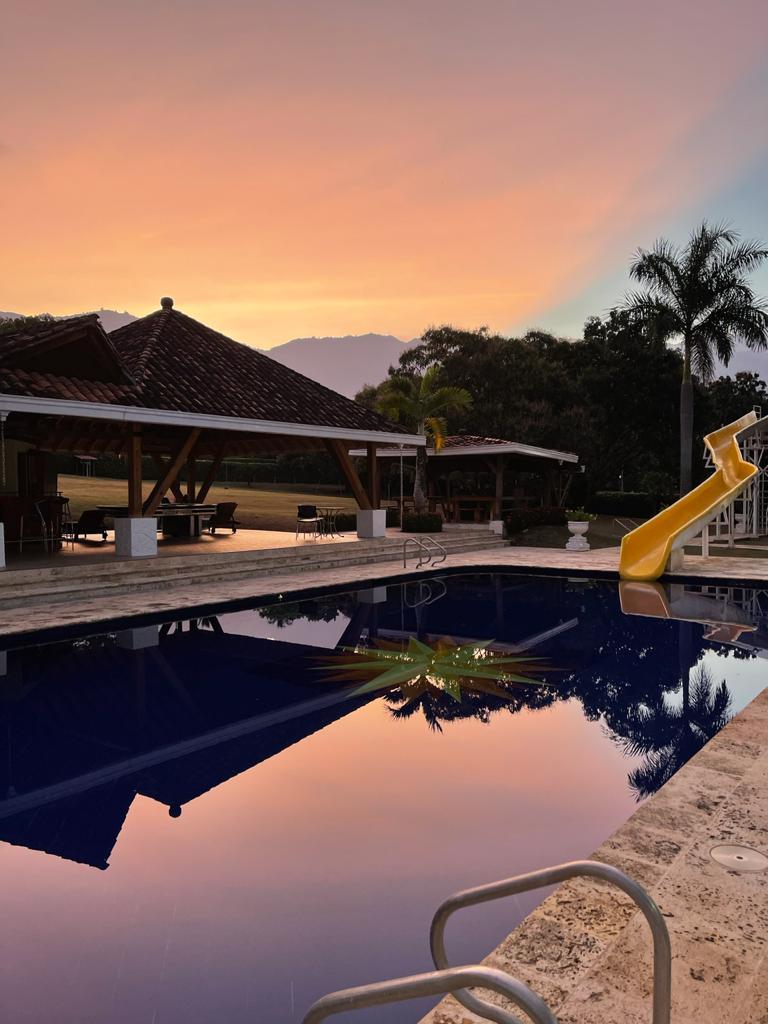 The image features a tranquil pool area reflecting a sunset sky, with a yellow slide positioned next to the pool. An open pavilion is visible in the background, surrounded by lush greenery, while lounge chairs are placed nearby, creating a serene outdoor setting.