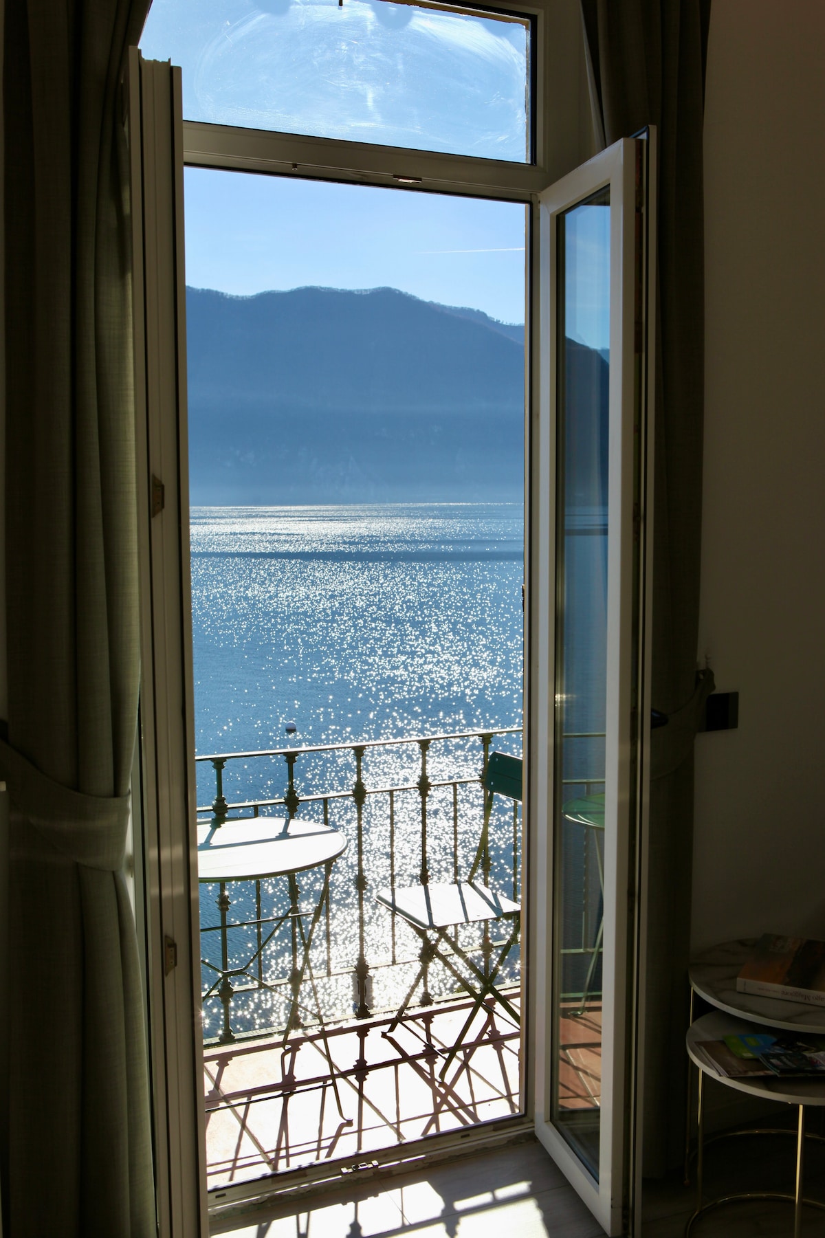 A set of open glass doors leads to a private balcony, offering an unobstructed view of shimmering lake waters and distant mountains. A small round table with two chairs is positioned on the balcony, inviting relaxation and enjoyment of the scenic surroundings.