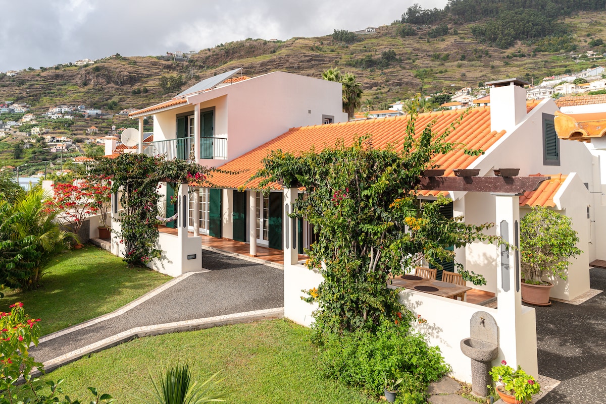 A two-story house is framed by vibrant greenery, featuring a traditional orange-tiled roof. The front porch showcases multiple large windows, allowing natural light to illuminate the inviting entrance. Flowering plants enhance the exterior, while gently sloped landscaping guides the eye towards the home.