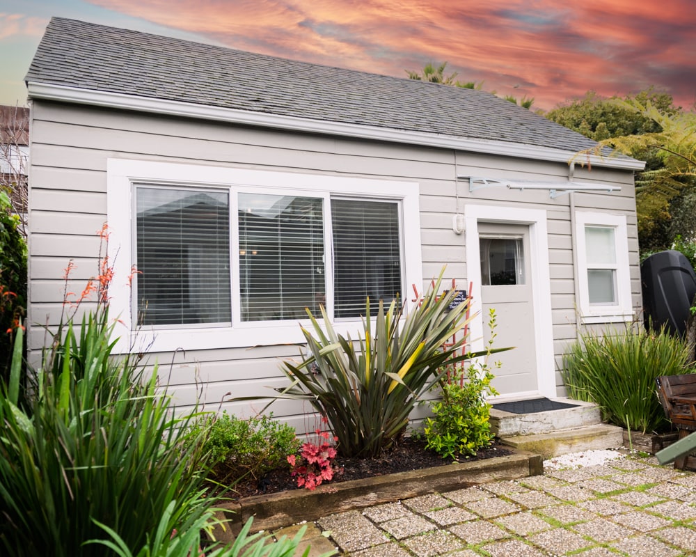 The exterior of a standalone cottage is presented, featuring a light gray siding and white trim. A welcoming entrance is framed by vibrant green plants and flowers. Large windows allow for natural light to enter, creating an inviting appearance against a colorful sunset sky.