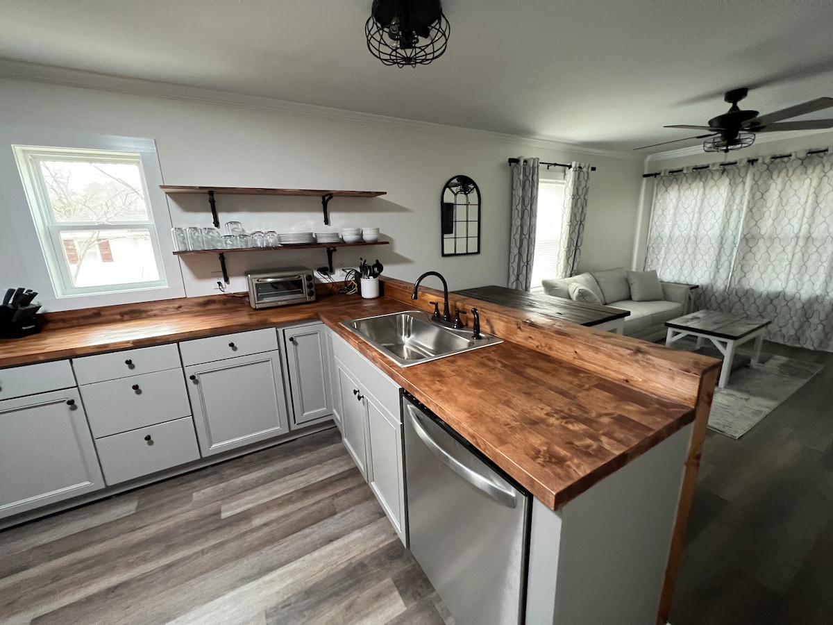 The kitchen features a combination of modern appliances and a wooden countertop, enhancing functionality. Open shelving holds dishware, while a window offers natural light. Adjacent seating is visible, with light-colored furniture and soft furnishings arranged for relaxation.