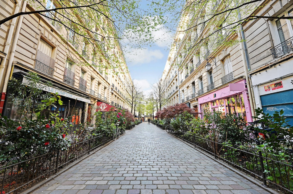 A tranquil pedestrian street lined with charming foliage and colorful storefronts. Smooth cobblestones create a welcoming path, framed by greenery that enhances the serene atmosphere. Sunlight filters through the trees, adding a warm glow to the inviting scene.