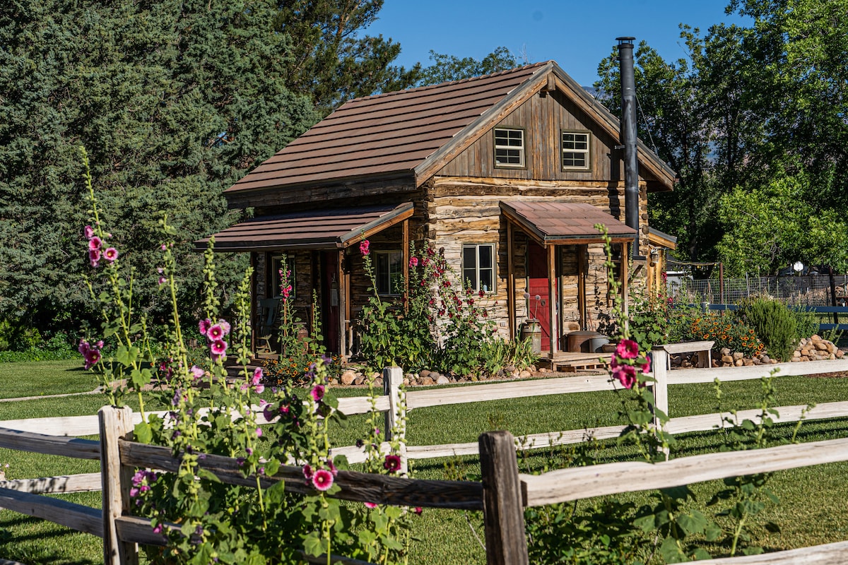 A rustic log cabin is framed by blooming flowers in the foreground and surrounded by lush greenery. The cabin features a metal roof, wooden walls, and a chimney, complemented by a wooden fence and a well-maintained lawn.