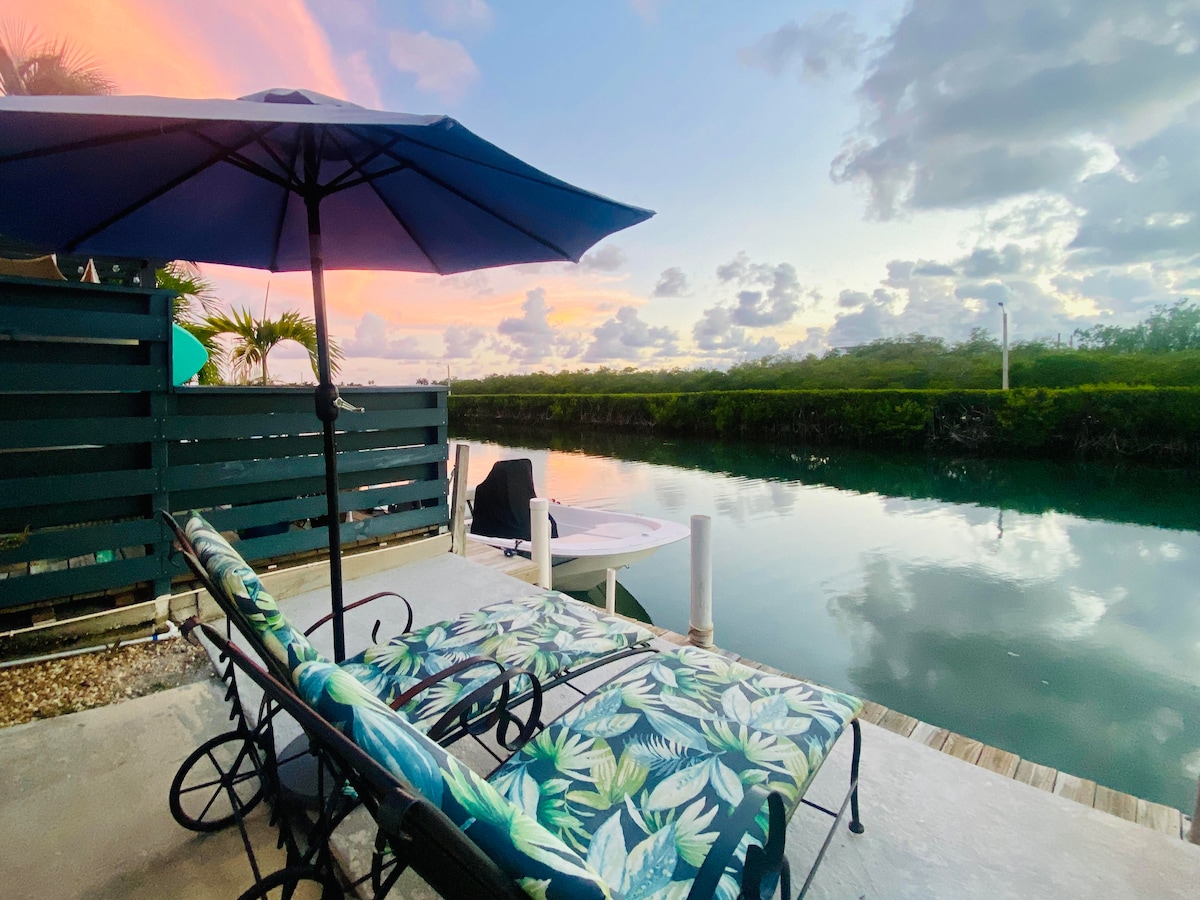 A waterfront patio is shown with two lounge chairs featuring tropical-patterned cushions, under a large blue umbrella. The calm water reflects the colorful sky at sunset, with lush greenery lining the canal for a scenic view.