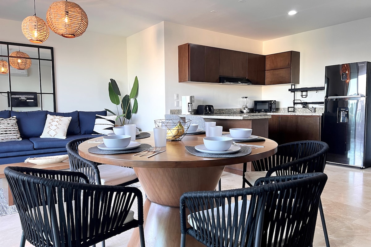 A dining area features a round wooden table surrounded by six black woven chairs. Five white bowls are placed neatly on the table, complemented by a fruit bowl in the center. Modern kitchen cabinetry and appliances are visible in the background, along with a cozy living space.