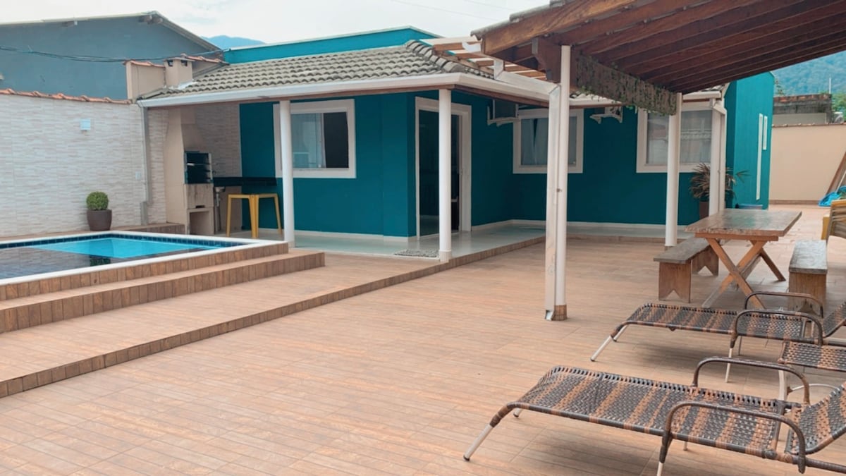 A spacious outdoor area features a refreshing pool, wooden decking, and a comfortable seating space. A covered patio with a picnic table is visible next to the house, providing shaded seating. The house exterior is painted in a calming blue, contributing to a relaxed atmosphere.