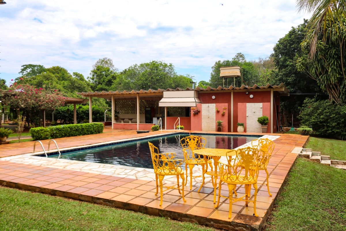 A rectangular pool is surrounded by a tiled patio, featuring yellow outdoor chairs and a small table. Lush greenery and flowering plants are visible in the background, alongside a structure that appears to contain amenities for guests.