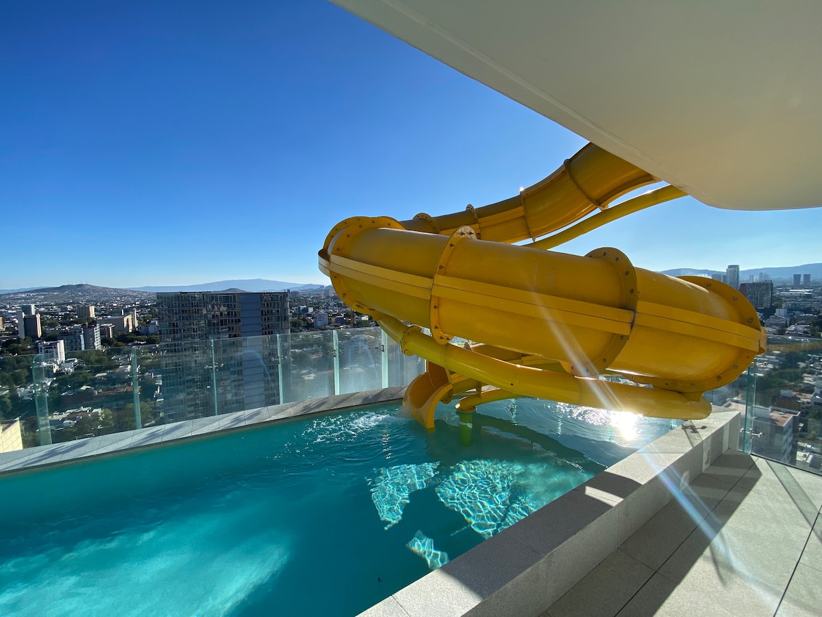 A vibrant yellow waterslide is positioned above a clear pool, offering a playful experience. The pool is surrounded by glass railings, providing an unobstructed view of the city skyline. Sunlight reflects off the water's surface, enhancing the inviting atmosphere of the rooftop area.