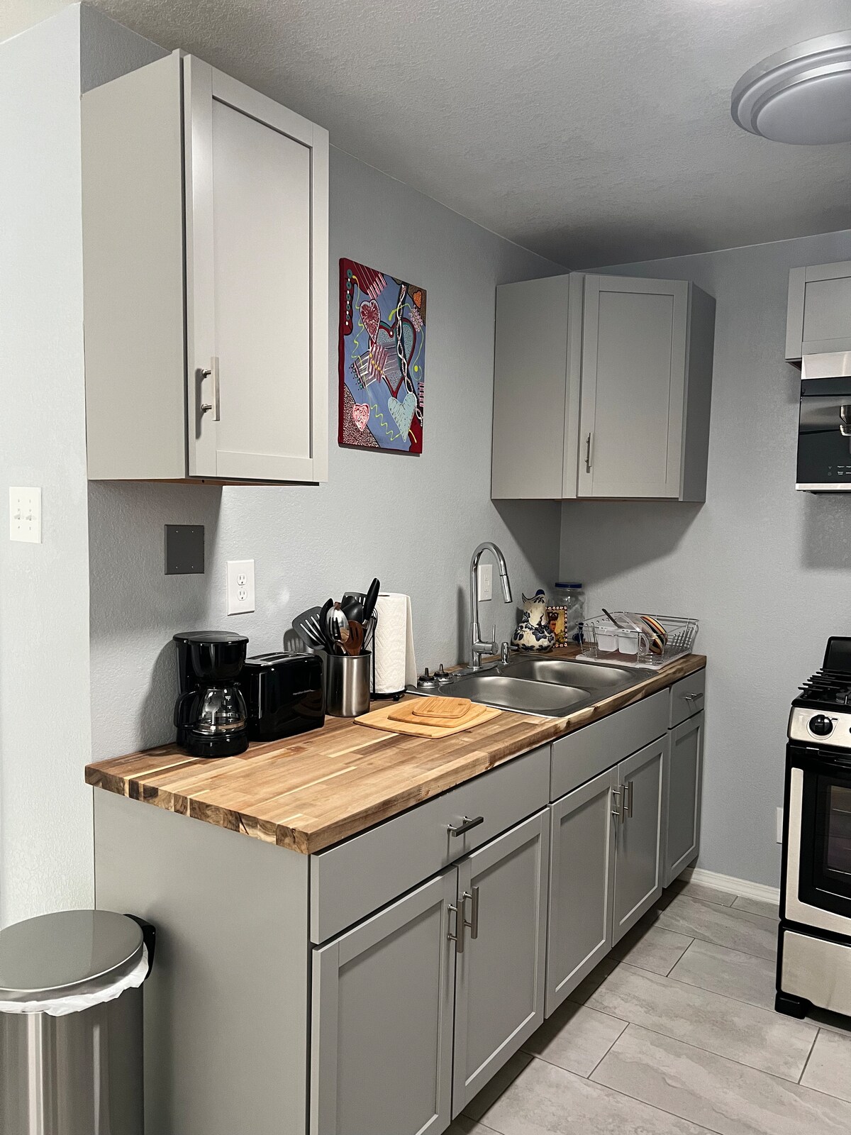 The compact kitchen features light grey cabinetry and a wooden countertop. A sink and stove are situated alongside essential appliances, including a coffee maker. A colorful piece of artwork adorns the wall, complementing the modern design.
