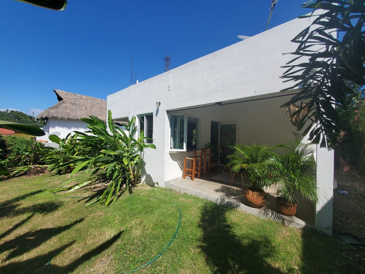 A white house is surrounded by lush greenery, featuring large windows that invite natural light. The exterior includes a small patio area with wooden chairs, framed by potted plants, creating a serene connection between indoor and outdoor spaces.