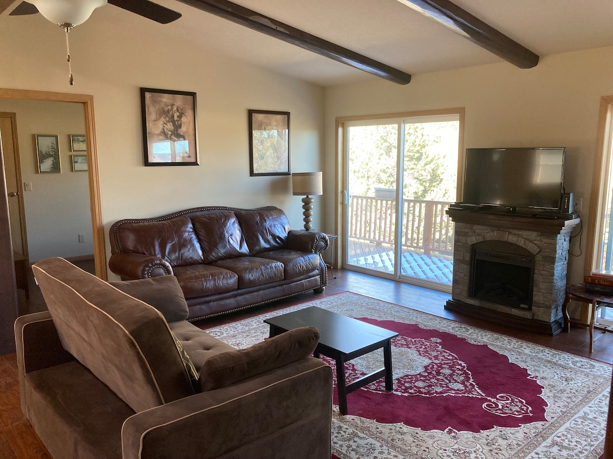 A comfortable living area is furnished with a brown leather sofa and an armchair, complemented by a small black coffee table. A stone fireplace adds warmth to the space. Large windows offer views of the outdoors, allowing natural light to fill the room.