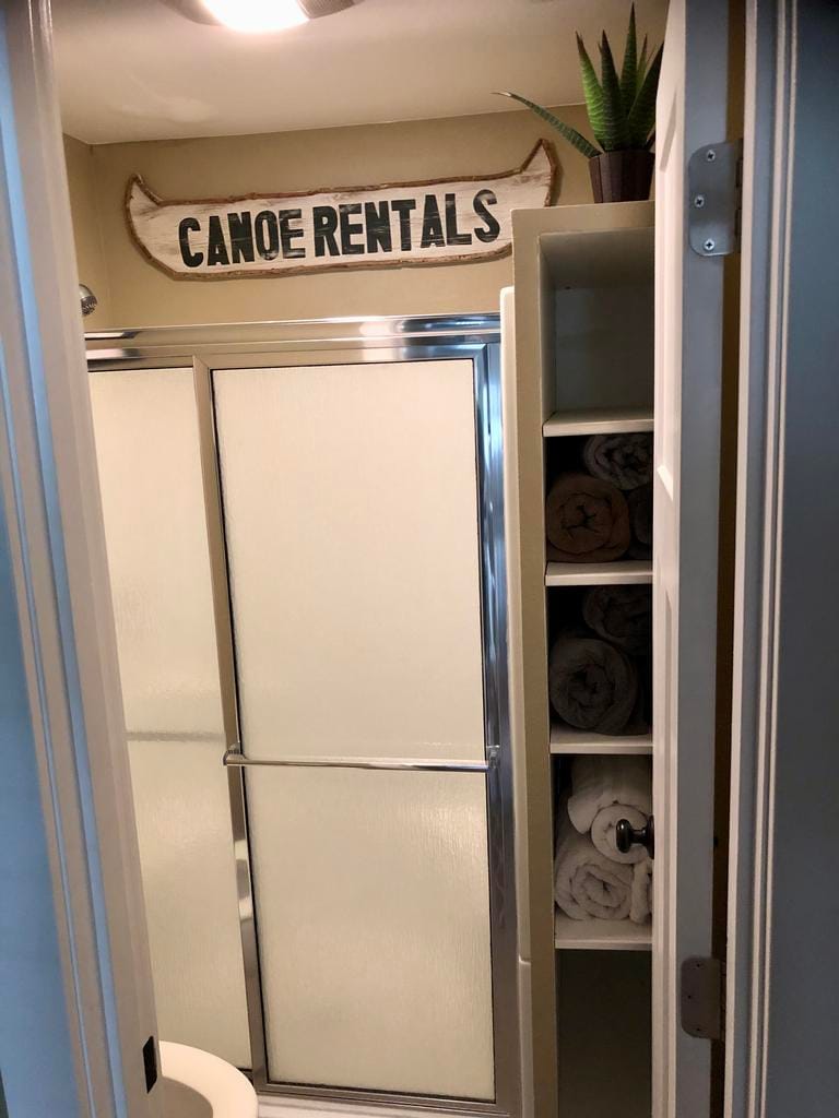A bathroom is shown featuring a glass shower enclosure and a toilet. Fresh towels are neatly arranged on a shelf adjacent to the shower. A sign reading 'Canoe Rentals' hangs above, complemented by a small plant positioned on top of the shelf.