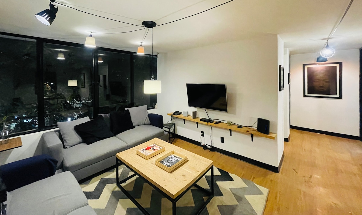 A modern living room contains a spacious gray sofa and a blue armchair, positioned around a wooden coffee table. A flat-screen TV is mounted on the wall, and soft lighting is provided by three pendant lamps. Large windows offer a view of the outdoor foliage.