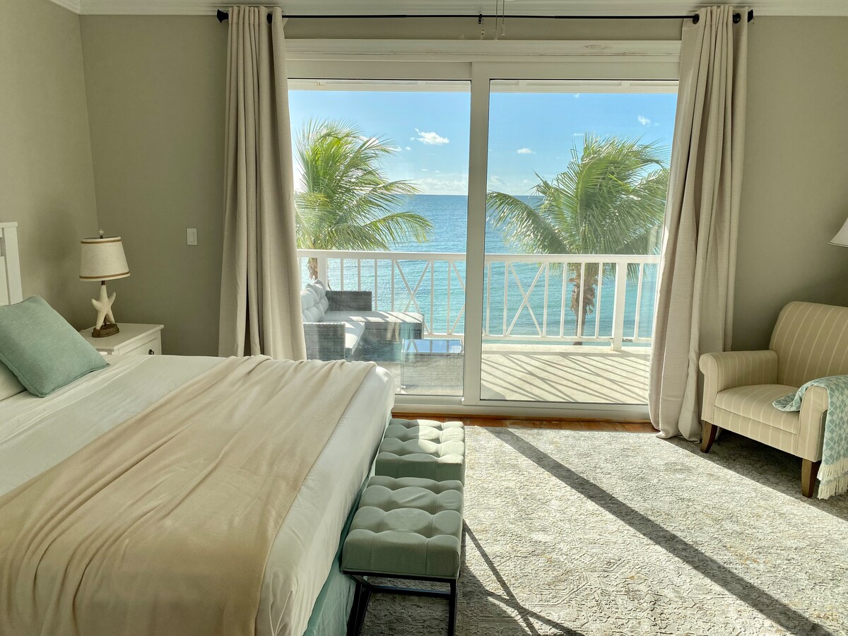 A serene bedroom is flooded with natural light from large sliding glass doors opening to a private terrace. Soft blue and beige tones are present, with a spacious bed adorned with neutral linens. A cozy armchair and a decorative bench add functional touches to the inviting space.