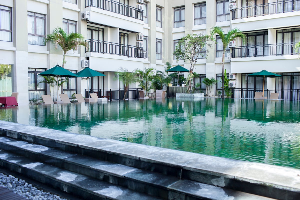 A serene swimming pool is surrounded by lounge chairs and umbrellas, presenting a calming reflection of the neighboring greenery. The pool area is flanked by the hotel’s modern architecture, with glass doors opening to the inner courtyard, fostering a tranquil atmosphere.