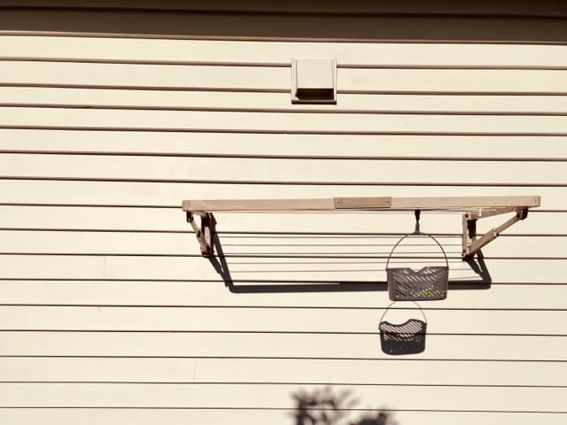 A simple wooden shelf is mounted on a beige exterior wall, supported by metal brackets. Below, a small basket hangs, providing additional storage. The shadows of the shelf and basket create a patterned effect on the wall beneath them.