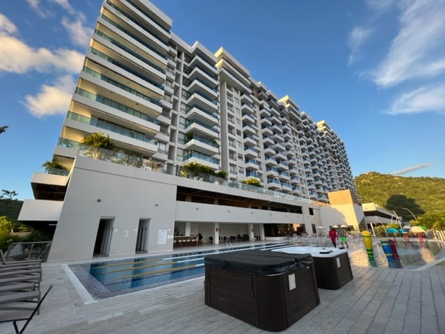 A modern multi-story building features several balconies with glass railings, overlooking a large pool area. An inviting swim-up bar is visible on the ground level, alongside a well-maintained landscaped courtyard. The backdrop includes lush green hills under a clear blue sky.