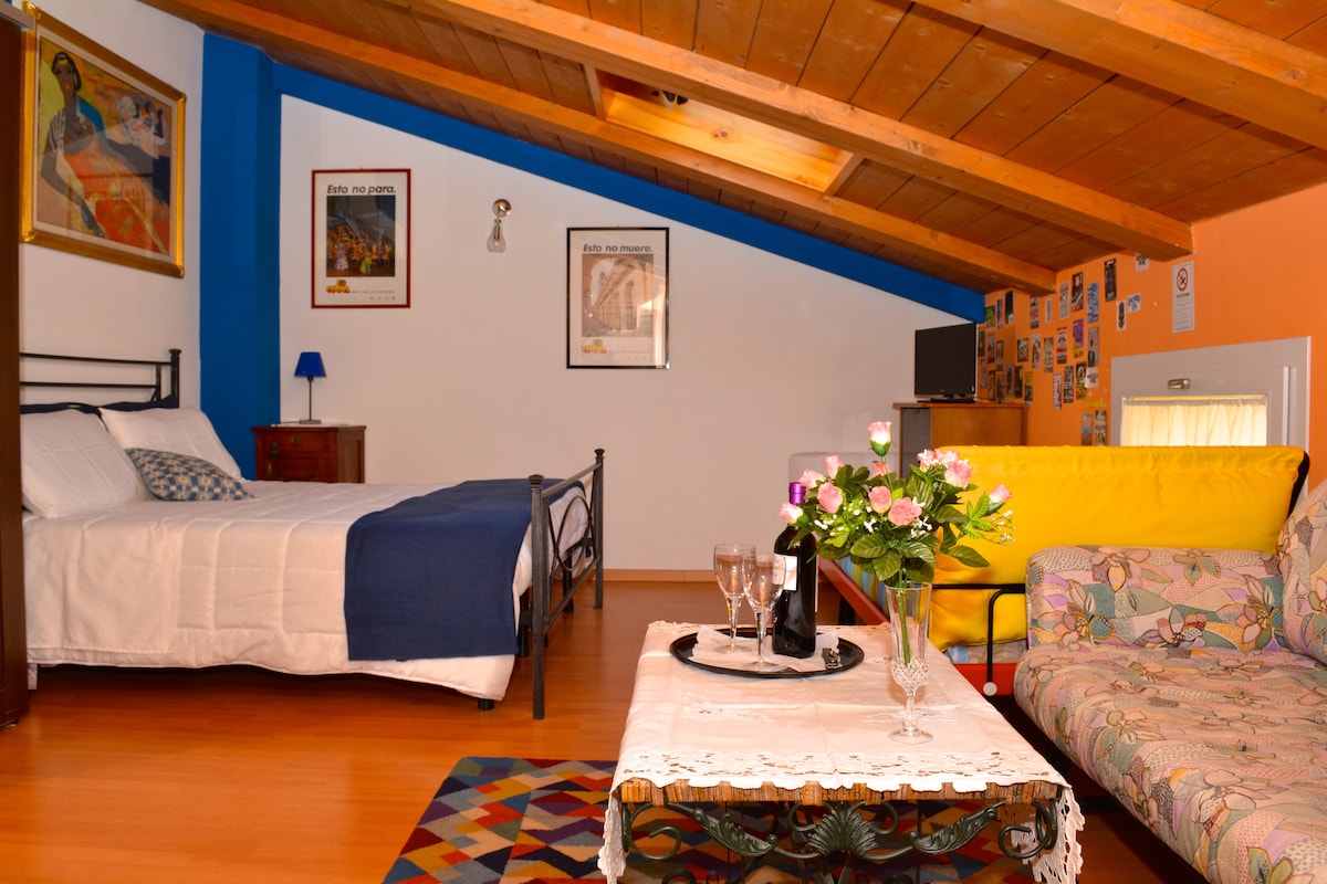 A spacious room features a double bed with a bright white coverlet and a blue accent wall. A cozy yellow sofa and a coffee table with a floral arrangement sit in the corner. Natural light illuminates the space from a window under a sloped wooden ceiling.