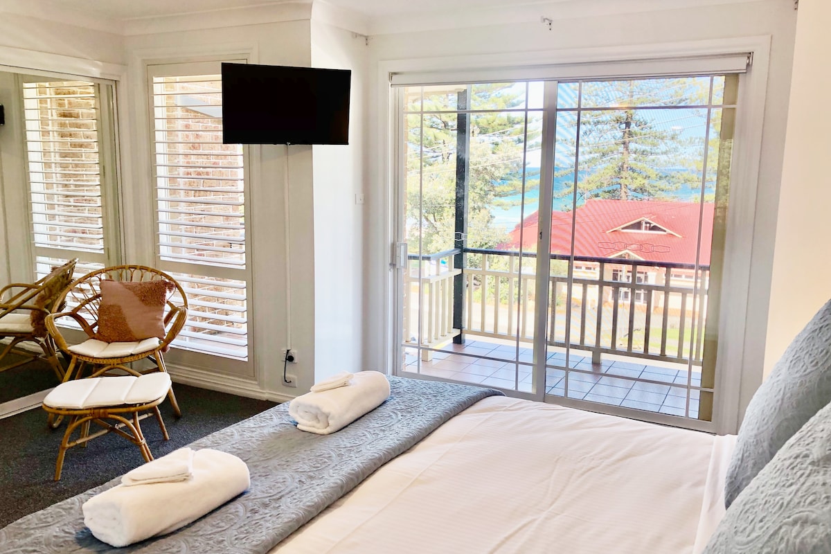 A bright bedroom features a large king-size bed with fresh linens and towels. Sunlight filters through the glass sliding doors, leading to a private balcony with views of the ocean. A minimalist seating area is provided with a chair and side table.