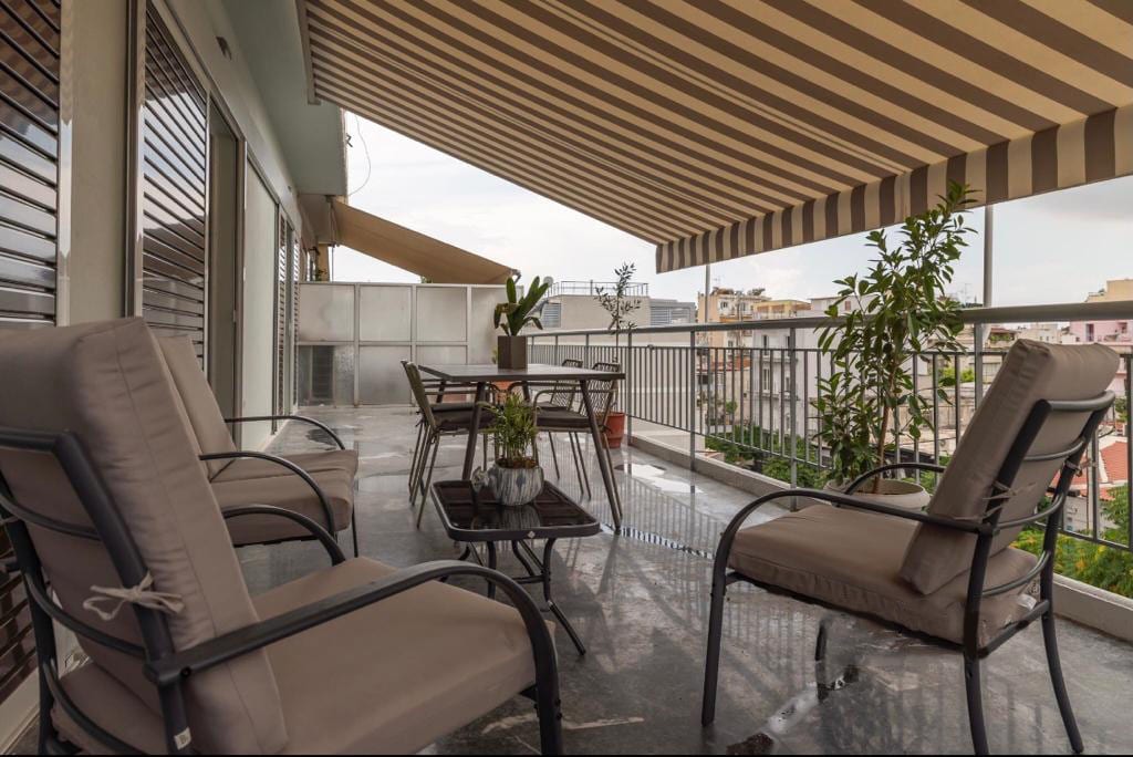 An extra large terrace is furnished with comfortable lounge chairs and a dining table surrounded by potted plants. A striped awning provides shade, and the polished flooring reflects the natural light, creating a relaxing outdoor space with views of the surrounding area.