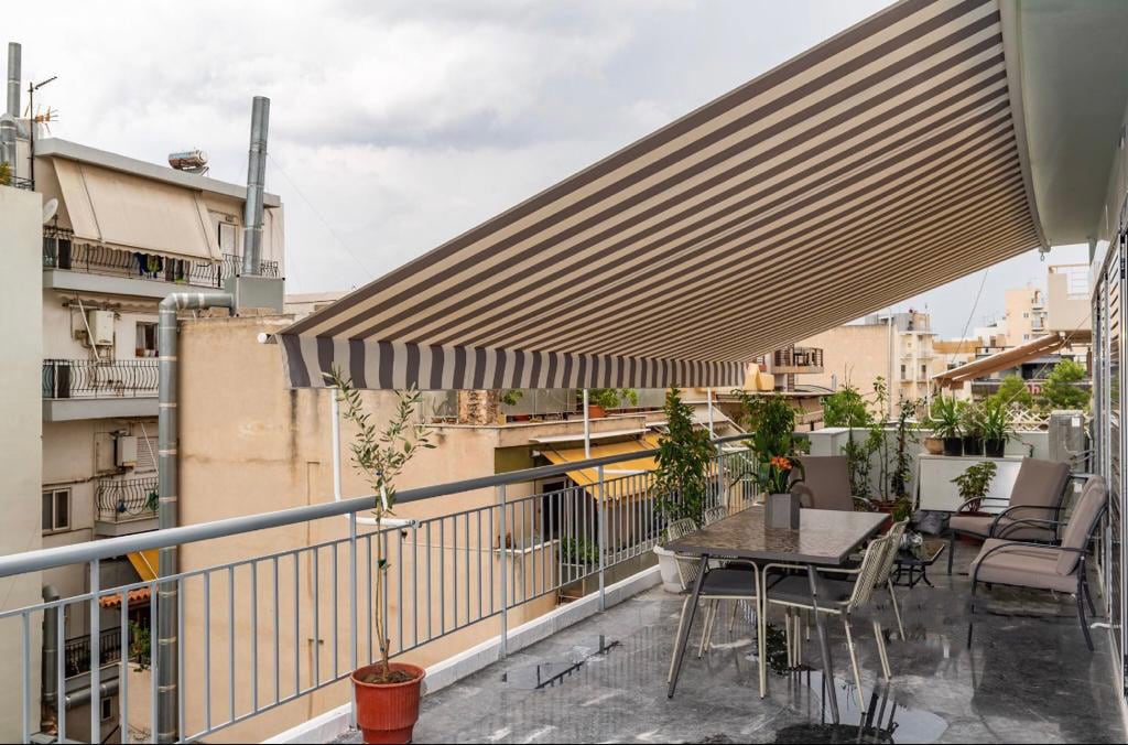 An extra large terrace is featured, equipped with a dining table and several lounge chairs, all under a striped awning. Potted plants add greenery to the space, while the surrounding buildings are visible beyond the railing.