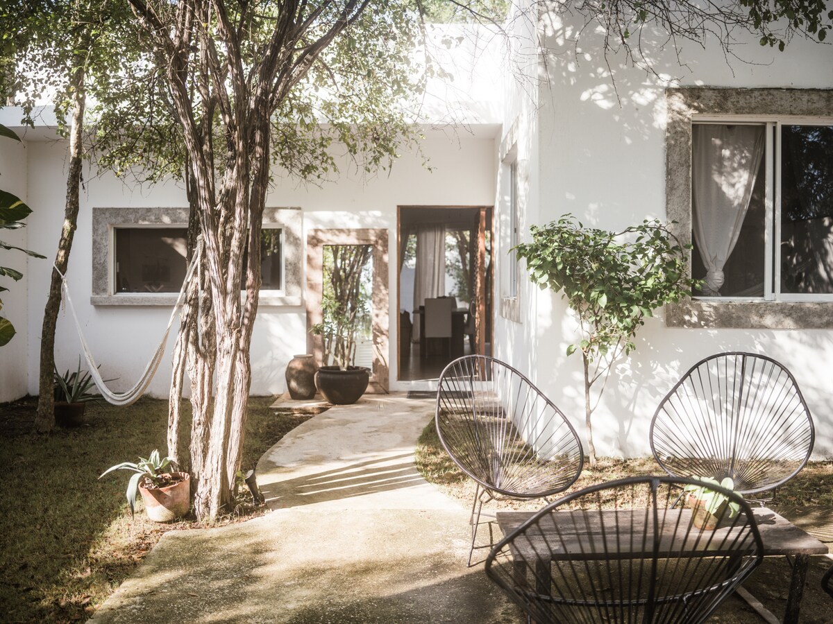 An inviting outdoor space features a winding path leading to a private entrance. Two black woven chairs sit on a sunlit patio surrounded by lush greenery and potted plants. A hammock hangs between trees, complemented by the textured walls of the home in the background.