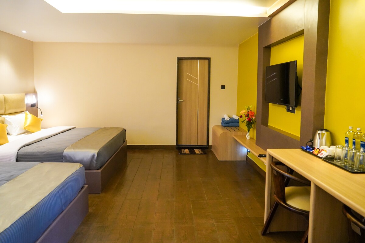 A spacious family room features two comfortable beds dressed in neutral linens. A wooden desk and chairs are positioned beside a welcoming beverage station. A vivid yellow accent wall brings warmth to the space, while a wall-mounted television adds modernity.