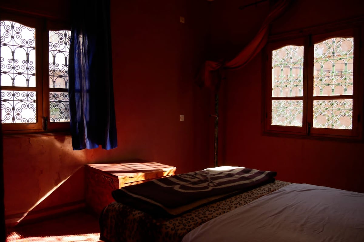 A warm-toned bedroom features a spacious bed with a layered blanket. Two ornate windows allow light to filter in, illuminating the textured walls. A wooden chest sits in the corner, enhancing the room's character with traditional design elements.
