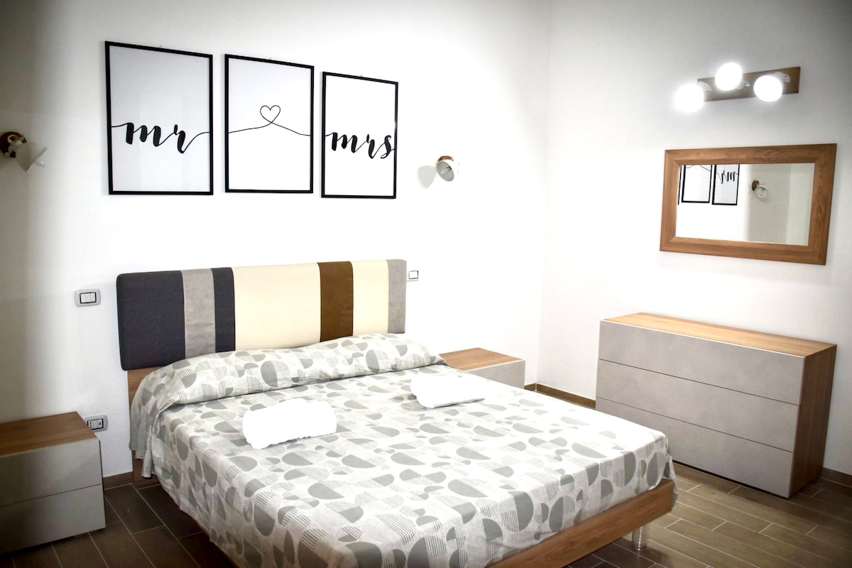 A spacious bedroom features a large bed with a patterned duvet and white towels neatly arranged. Modern decor includes framed artwork on the walls and a mirror above a minimalist dresser. Soft lighting is provided by a wall fixture and overhead lights.