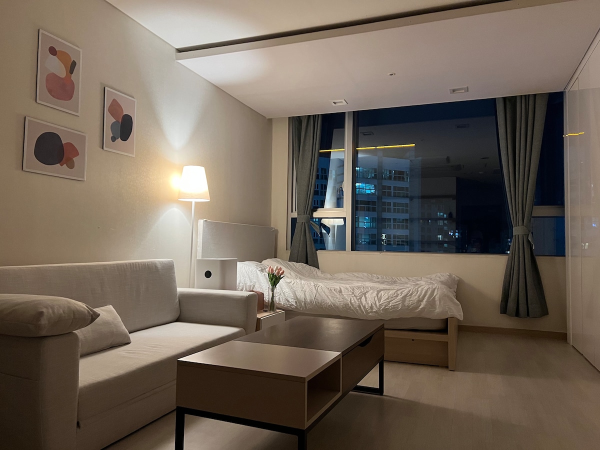 A bright living space is offered, featuring a cozy sofa and a coffee table. A spacious bed is visible in the background, with soft bedding arranged neatly. Large windows allow for ample natural light, and decorative wall art adds a subtle touch.