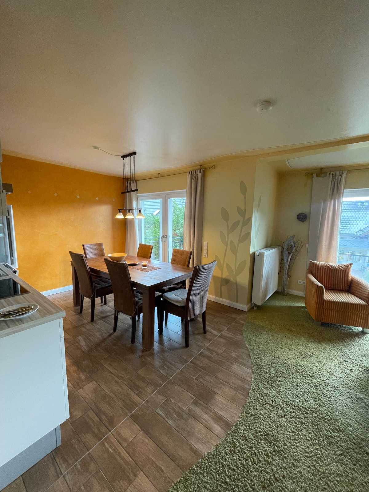 A dining area features a wooden table surrounded by six chairs, with warm yellow walls adding a cheerful touch. Soft lighting hangs above the table, and large windows allow natural light to enter, overlooking a garden. A cozy seating area is positioned in the corner.