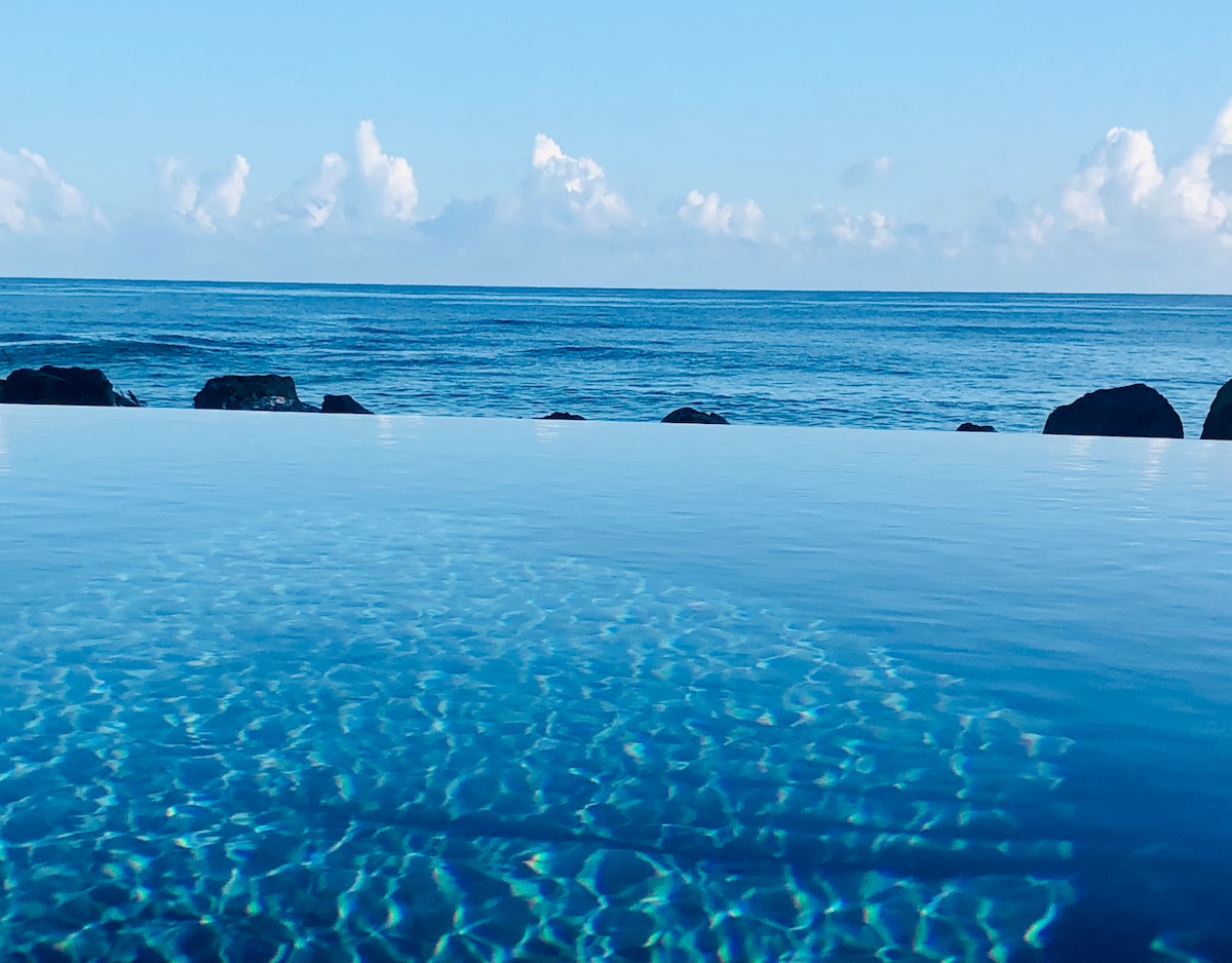 An infinity pool extends towards the horizon, merging with the ocean's calm waters under a clear blue sky. The surface reflects gentle ripples, while smooth rocks outline the pool's edge, creating a serene atmosphere.