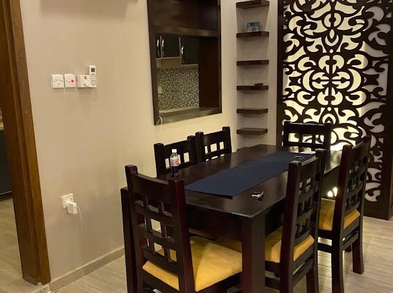 A dining area is depicted with a dark wooden table surrounded by six chairs featuring yellow cushions. An intricate decorative screen adds texture to the space, complemented by shelves holding minimal decorative items. A small kitchen is visible in the background.