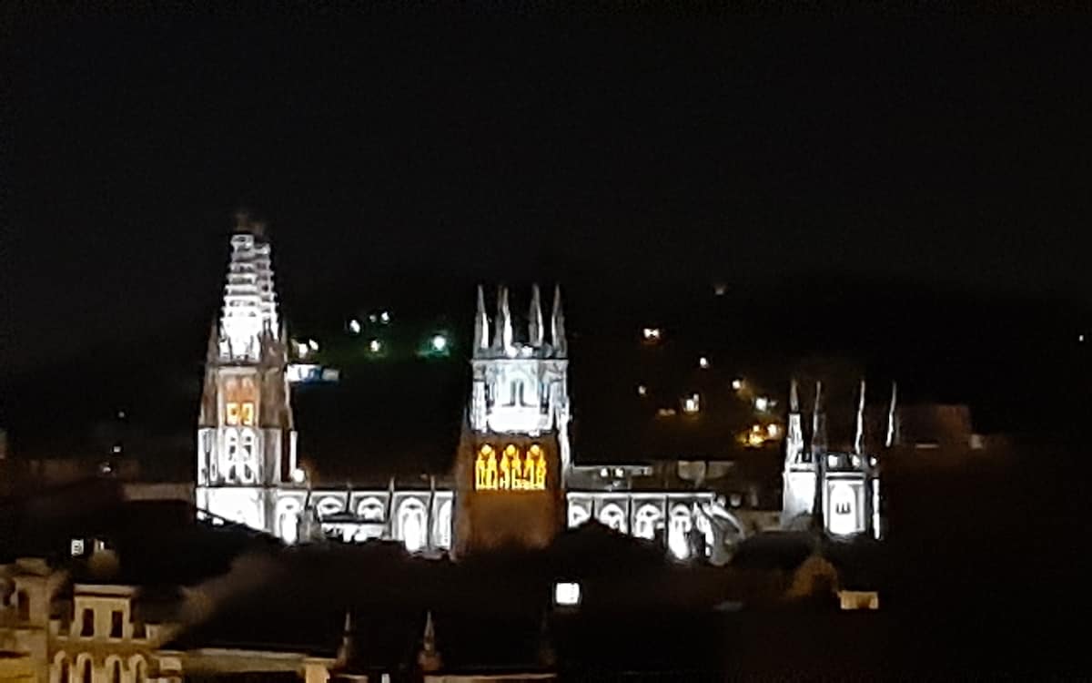 An illuminated view of a historic cathedral is displayed at night, showcasing its intricate architectural details. The facade is highlighted by soft lighting, revealing the towers and stained glass windows. Surrounding city lights create a scenic backdrop against the dark sky.
