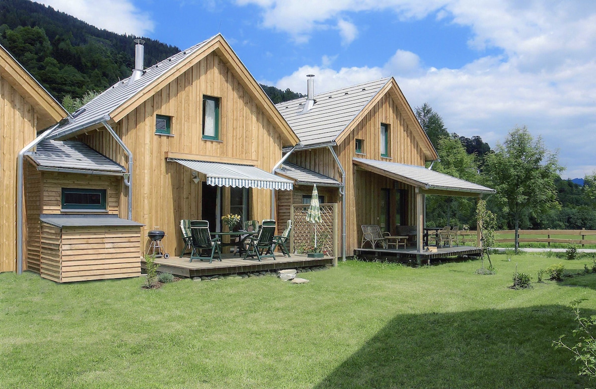 A wooden house is set against a backdrop of lush hills, featuring a green lawn and outdoor seating area. A shaded terrace is visible, equipped with chairs and a table. Adjacent structures show a similar architectural style, contributing to a cohesive aesthetic.