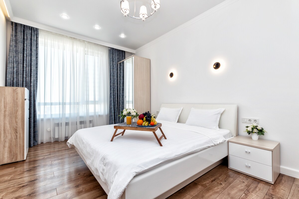 A well-lit bedroom features a spacious bed with white linens, complemented by a decorative tray of fresh fruit. Nearby, a mirrored wardrobe and a bedside table are visible, while large windows are adorned with sheer curtains, allowing natural light to fill the room.