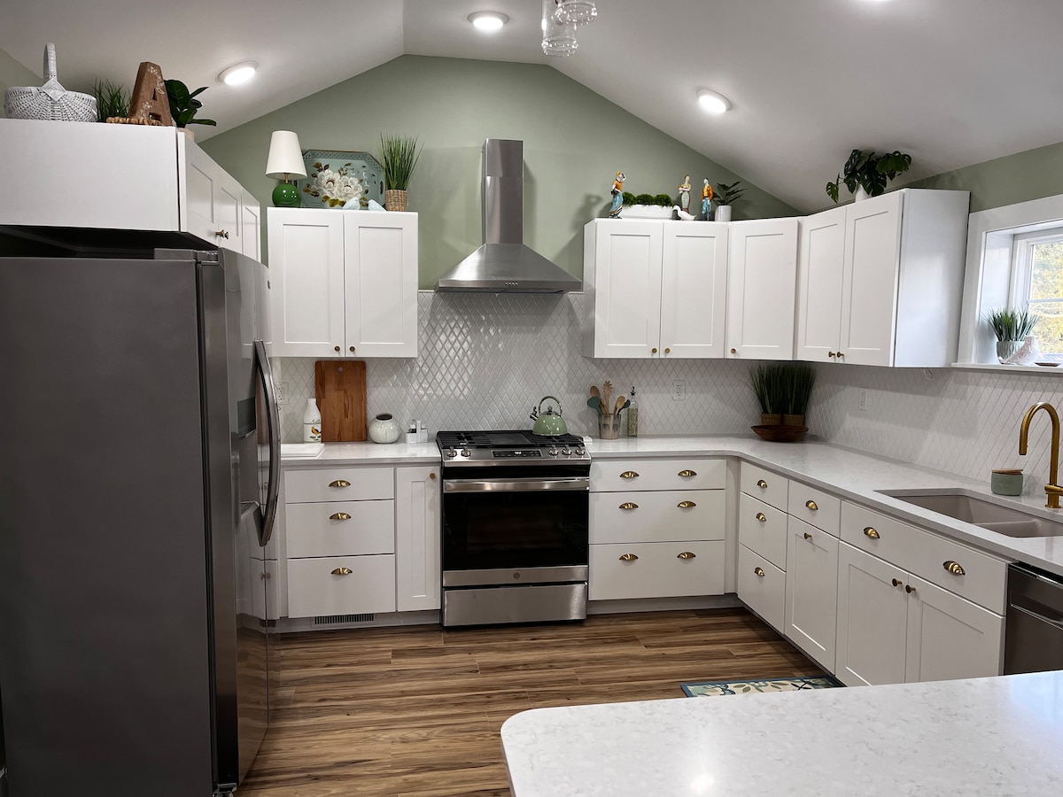 A modern kitchen features white cabinetry with brass handles, complemented by a stainless steel gas stove and range hood. Natural light fills the space through a window above the sink, enhancing the neutral color scheme of light greens and whites, with decorative plants accenting the countertops.