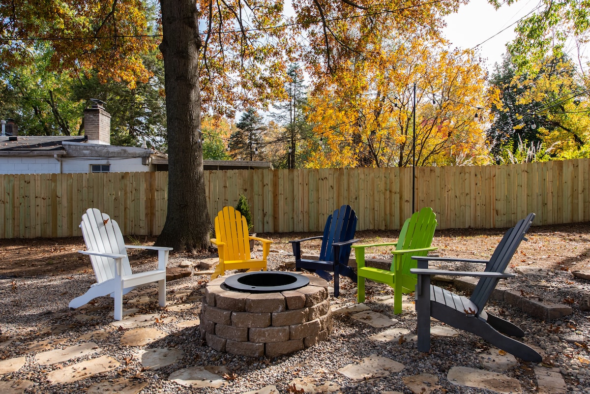 A fire pit surrounded by colorful Adirondack chairs stands in a gravel area, framed by a wooden fence and trees displaying autumn foliage. The seating arrangement invites relaxation, making it an ideal spot for gatherings.