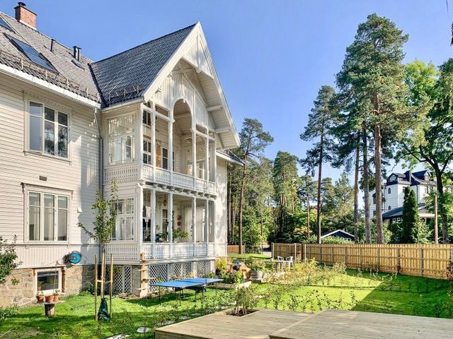 A charming Swiss villa features a large balcony with decorative railing, surrounded by tall trees. The spacious garden is well-maintained, with several seating areas and greenery. The facade showcases elegant architectural details, adding to the overall character of the property.