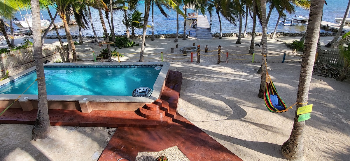The image captures a spacious outdoor area featuring a pool surrounded by palm trees. A hammock is visible nearby, along with a sandy beach leading to the water. A private pier is located in the background, with boats moored along the shore.