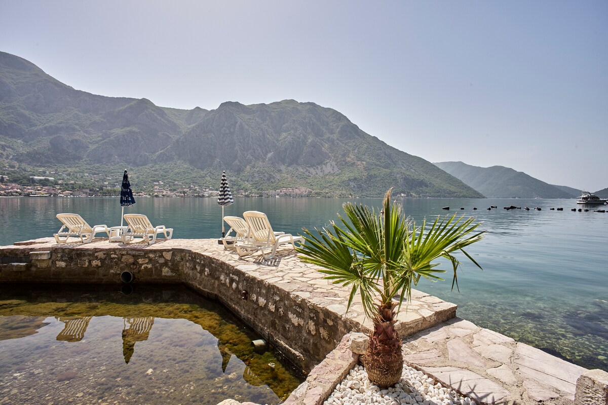 A stone jetty extends into calm, clear waters, surrounded by lush mountains. Several beach chairs are positioned along the edge, accompanied by a large umbrella and a small palm tree. The tranquil setting reflects the serene atmosphere of a seaside retreat.